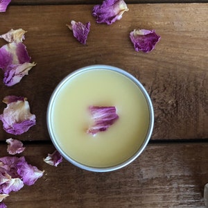 Puede incluir: Un pequeño recipiente redondo de bálsamo amarillo pálido con un solo pétalo de rosa seco en la parte superior. El recipiente está sobre una superficie de madera con pétalos de rosa secos esparcidos alrededor.