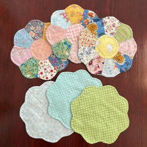 May include: Five flower-shaped fabric coasters. Three coasters are made of patchwork fabric with different floral patterns. Two coasters are solid colours with white polka dots on a light blue and light green background.