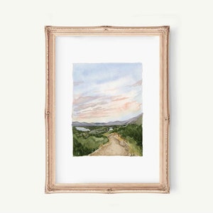 May include: Watercolor painting of a landscape with a dirt road leading up a hill towards a mountain range. The sky is a soft blue with pink clouds. The painting is framed in a light wood frame.