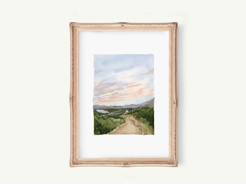May include: Watercolor painting of a landscape with a dirt road leading up a hill towards a mountain range. The sky is a soft blue with pink clouds. The painting is framed in a light wood frame.