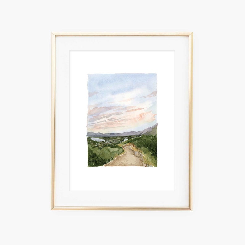 May include: A watercolor painting of a winding dirt road leading up a hill towards a mountain range. The sky is a soft blue with pink clouds. The painting is framed in a gold frame.