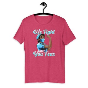 May include: Pink t-shirt with a graphic of a woman wearing a blue surgical mask and scrubs. The text "We Fight What You Fear" is printed on the shirt.