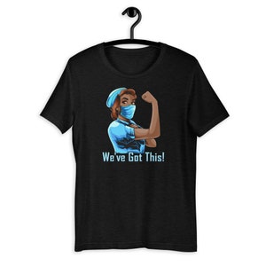 May include: Black t-shirt featuring a graphic of a woman in a medical mask and cap flexing her arm. The image is in a comic book style with the text "We've Got This!" in blue below the image.