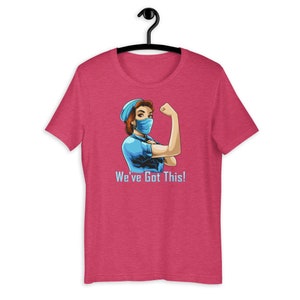 May include: A short-sleeve, pink t-shirt featuring a cartoon illustration of a woman in a blue medical mask and cap flexing her arm. The text "We've Got This!" is printed below the image in light blue.