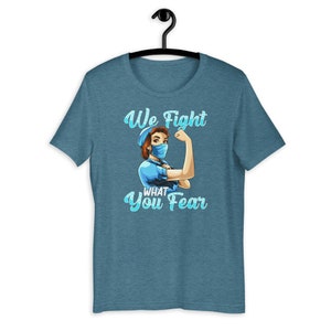 May include: A teal blue t-shirt with a graphic of a woman wearing a blue face mask and a blue shirt with the text "We Fight What You Fear".