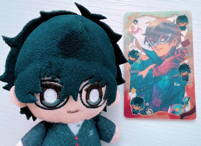 Puede incluir: Un peluche de un personaje con cabello negro, ojos verdes y un atuendo verde y blanco. El peluche sostiene una tarjeta con una ilustraci&oacute;n colorida de un personaje con una m&aacute;scara y un atuendo rojo y azul.