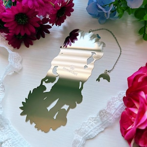 May include: A silver-toned metal bookmark with a reflective surface featuring a die-cut silhouette of a figure. The bookmark includes a thin silver chain attached to a small dolphin charm. The text "SAVERIO ARTS" is engraved on the center surface.