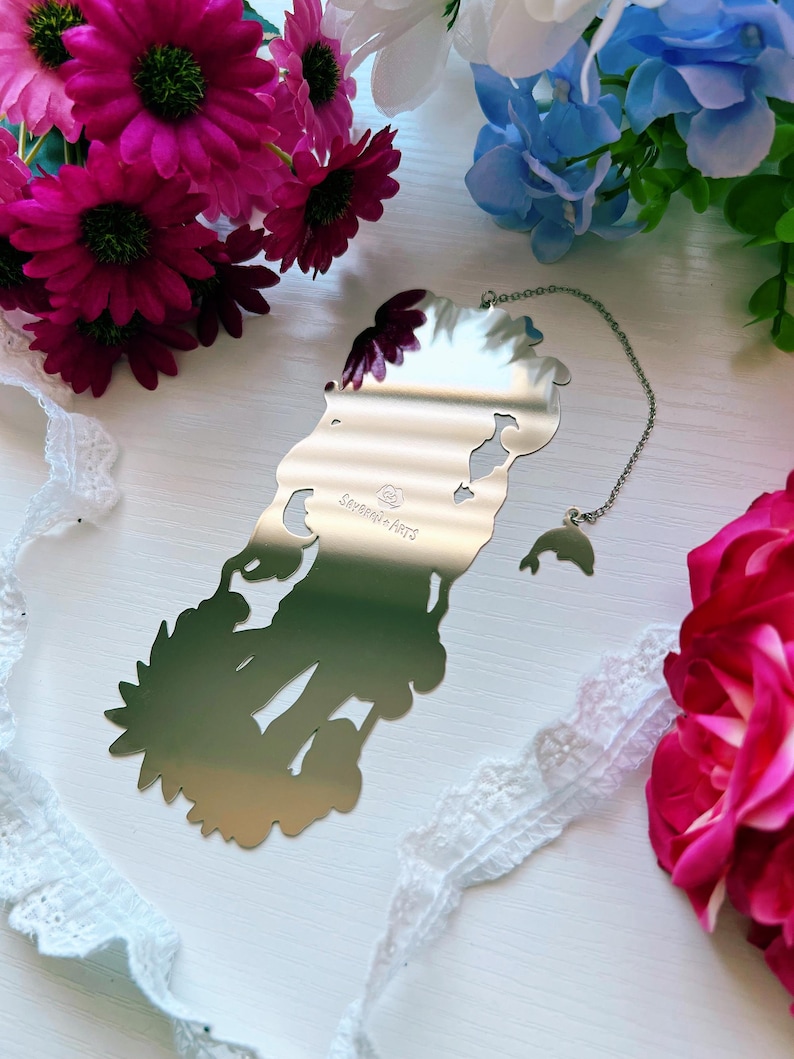 May include: A silver-toned metal bookmark with a reflective surface featuring a die-cut silhouette of a figure. The bookmark includes a thin silver chain attached to a small dolphin charm. The text "SAVERIO ARTS" is engraved on the center surface.