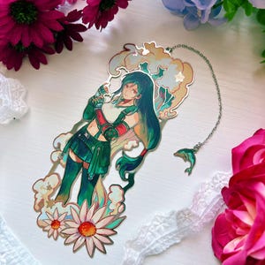 May include: A metal bookmark featuring a long-haired character in a green outfit, surrounded by stylized clouds and flowers. The bookmark has a silver chain attached with a small dolphin charm. The design includes soft pastel colors and intricate outlines.