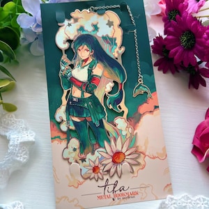 May include: A metal bookmark featuring an anime-style character with long dark hair, wearing a white top and green skirt, surrounded by stylized clouds and flowers. A thin silver chain with a small dolphin charm is attached to the top of the bookmark.