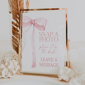 Può includere: Un cartello con cornice dorata con un fiocco rosa ad acquerello e il testo "SNAP A PHOTO place it in the book LEAVE A MESSAGE".
