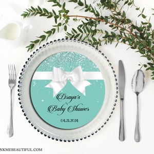 Puede incluir: Un plato redondo de papel azul turquesa con una cinta blanca y un lazo, purpurina plateada y texto negro que dice "Draya's Baby Shower 04.21.2024".