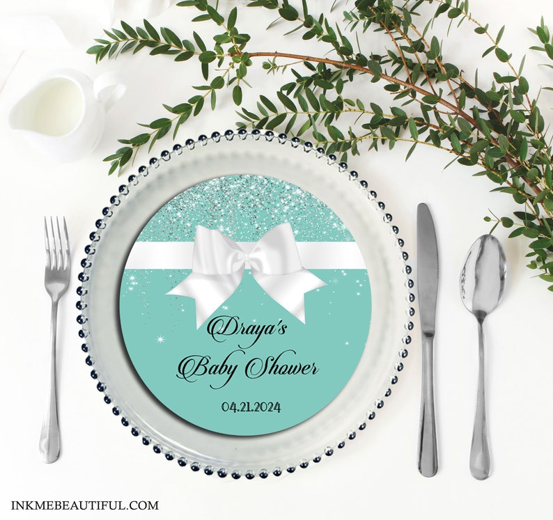 Puede incluir: Un plato redondo de papel azul turquesa con una cinta blanca y un lazo, purpurina plateada y texto negro que dice "Draya's Baby Shower 04.21.2024".