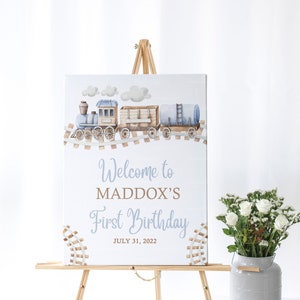 May include: A blue and brown train with white clouds on a light blue background. The sign reads "Welcome to Maddox's First Birthday July 31, 2022".
