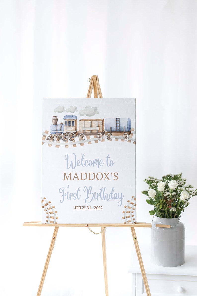 May include: A blue and brown train with white clouds on a light blue background. The sign reads "Welcome to Maddox's First Birthday July 31, 2022".
