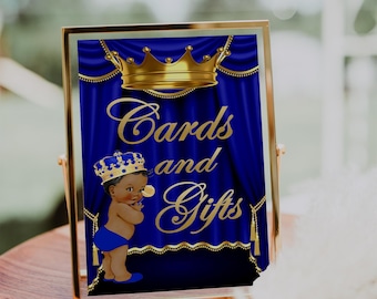 Puede incluir: Un cartel con marco dorado, fondo azul y texto dorado que dice "Tarjetas y Regalos". Un bebé de dibujos animados con corona dorada y pañal azul está en el lado izquierdo del cartel.