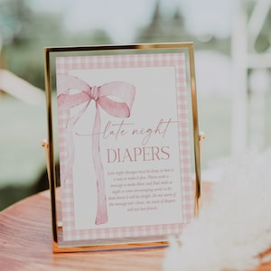 May include: A framed sign with a pink gingham pattern and a watercolor bow. The sign reads "late night DIAPERS" with additional text below. The frame is gold and the sign is displayed on a wooden surface.