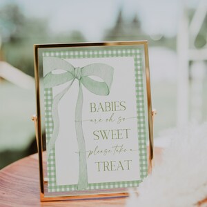 May include: A gold-framed sign with a green gingham background and a green watercolor bow. The sign reads "Babies are oh so sweet please take a treat."
