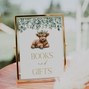 May include: A gold framed sign with a watercolor illustration of a brown Highland cow wearing a flower crown. The sign says "BOOKS and GIFTS" in gold lettering.