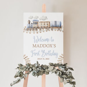 May include: A white sign with a blue and brown train illustration, the text "Welcome to Maddox's First Birthday July 31, 2022" and a decorative garland of green leaves.