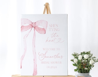 May include: A white canvas sign with a pink watercolor bow and the text "She's tying the knot" in pink script. Below the bow is the text "Welcome to Samantha's Bridal Shower 06.29.24".