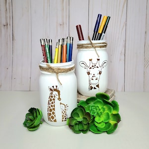 May include: Two white mason jars with gold giraffe designs. The jars are filled with pencils and markers. The jars are decorated with twine and are sitting on a white surface with green succulents.
