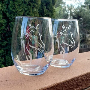 May include: Two clear glass wine glasses with etched horse head designs. The glasses are sitting on a wooden surface.