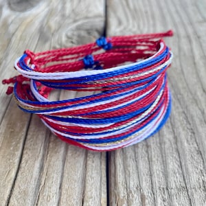 May include: A stack of red, white, blue, and gold string bracelets. The bracelets are made of tightly woven threads, with a mix of solid colors and metallic gold strands. The bracelets are displayed on a weathered wooden surface.
