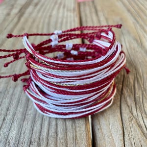 May include: A stack of handmade bracelets in red, white, and gold. The bracelets are made of woven cord and are stacked together. The bracelets are on a wooden surface.
