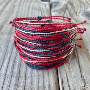 May include: A stack of braided bracelets in red, black, gold, and white. The bracelets are made of a thin, cord-like material and are tied with knots. The bracelets are stacked on a weathered wooden surface.