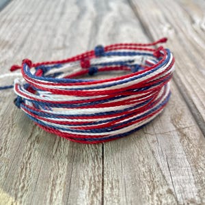 May include: A stack of patriotic-themed bracelets. The bracelets are made of red, white, and blue cords. The cords are tightly woven together, creating a layered effect. The bracelets are resting on a weathered wooden surface.