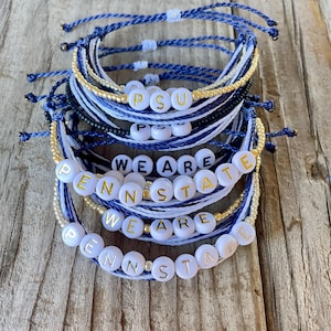 May include: Stack of handmade bracelets in shades of blue, white, and gold. The bracelets feature white letter beads spelling out "PSU" and "WE ARE PENN STATE". The bracelets are on a wooden surface.