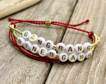 Könnte beinhalten: Ein handgefertigtes Armband mit weißen Buchstabenperlen, die "BANG NINER GANG" in goldenen Buchstaben buchstabieren. Das Armband hat rote und goldene Schnüre mit einem goldenen Perlenakzent. Das Armband liegt auf einer Holzoberfläche.