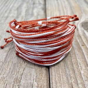 May include: A stack of handmade bracelets in shades of orange and white. The bracelets are made of woven cord and are stacked on top of each other. The bracelets are on a wooden surface.