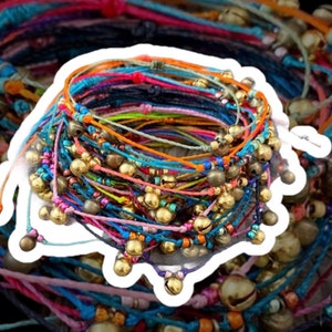 May include: A stack of colorful friendship bracelets with small gold-colored bells. The bracelets are made of various colored threads, including blue, orange, pink, and green. The bells are evenly spaced along the bracelets.