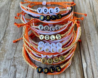 Peut inclure: Plusieurs bracelets faits à la main en orange, blanc, or et noir. Chaque bracelet affiche le mot "VOLS" avec des perles de lettres. Les bracelets sont faits de cordon et de perles, et sont probablement des produits dérivés.