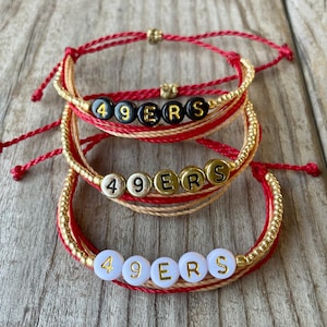May include: Three handmade bracelets with the text "49ERS" spelled out in letter beads. The bracelets feature red, gold, and tan cords, with gold-colored beads. The bracelets are displayed on a wooden surface.