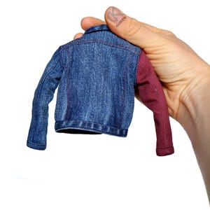 May include: A small denim jacket with one burgundy sleeve. The jacket is held by a hand.