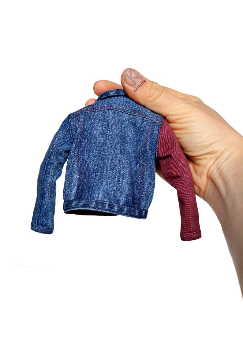 May include: A small denim jacket with one burgundy sleeve. The jacket is held by a hand.