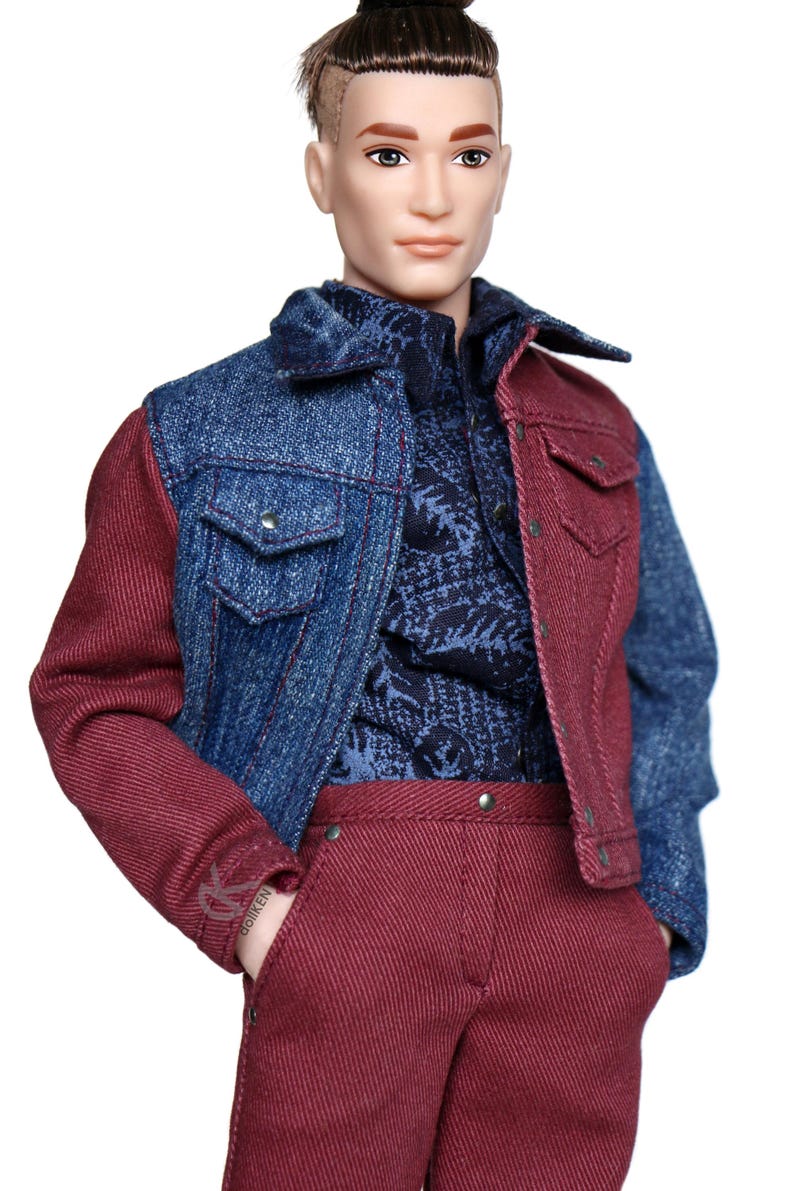 May include: A doll wearing a denim jacket with one red sleeve and one blue sleeve. The doll is also wearing a blue patterned shirt and red pants.