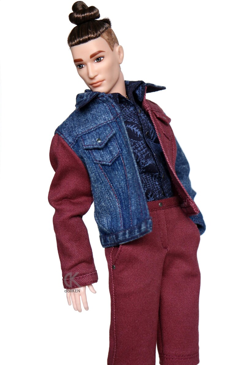 May include: A doll wearing a denim jacket with one red sleeve and red pants. The doll has brown hair in a bun and a shaved side.