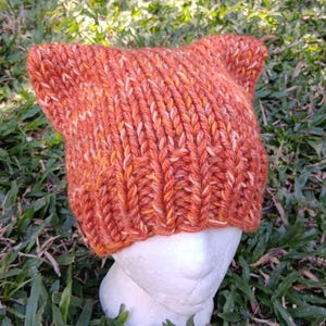 May include: A chunky knit beanie in a variegated orange and yellow yarn. The hat features a ribbed brim and two pointed ears at the top, creating a cat-like silhouette. The texture is thick and tactile, showcasing a prominent vertical stitch pattern.