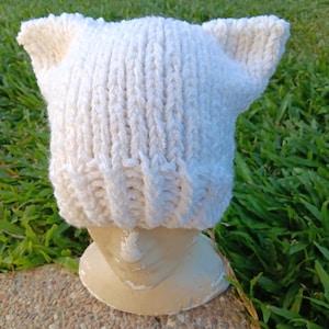 May include: White knitted hat with cat ears. The hat is made from a soft, fluffy yarn and has a simple, classic design.