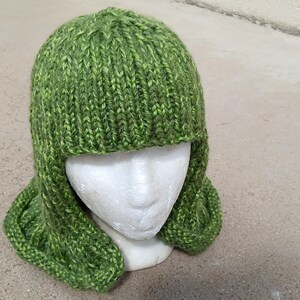 May include: A hand-knitted green beanie with ear flaps, crafted from variegated yarn in shades of lime and forest green. The hat features a ribbed texture throughout and is displayed on a white mannequin head, covering the ears and sides of the face.