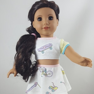 May include: A doll wearing a white crop top and skirt set with pastel skateboard and roller skate graphics. The top has yellow and blue accents. The doll has brown hair and is wearing purple sneakers.