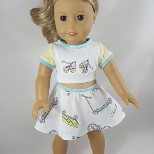 May include: A doll wearing a white crop top and skirt set with a skating theme. The top has yellow sleeves and blue trim, with roller skate graphics. The skirt features skateboards and scooters. The doll is wearing white sneakers.