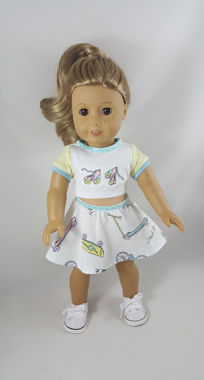 May include: A doll wearing a white crop top and skirt set with a skating theme. The top has yellow sleeves and blue trim, with roller skate graphics. The skirt features skateboards and scooters. The doll is wearing white sneakers.