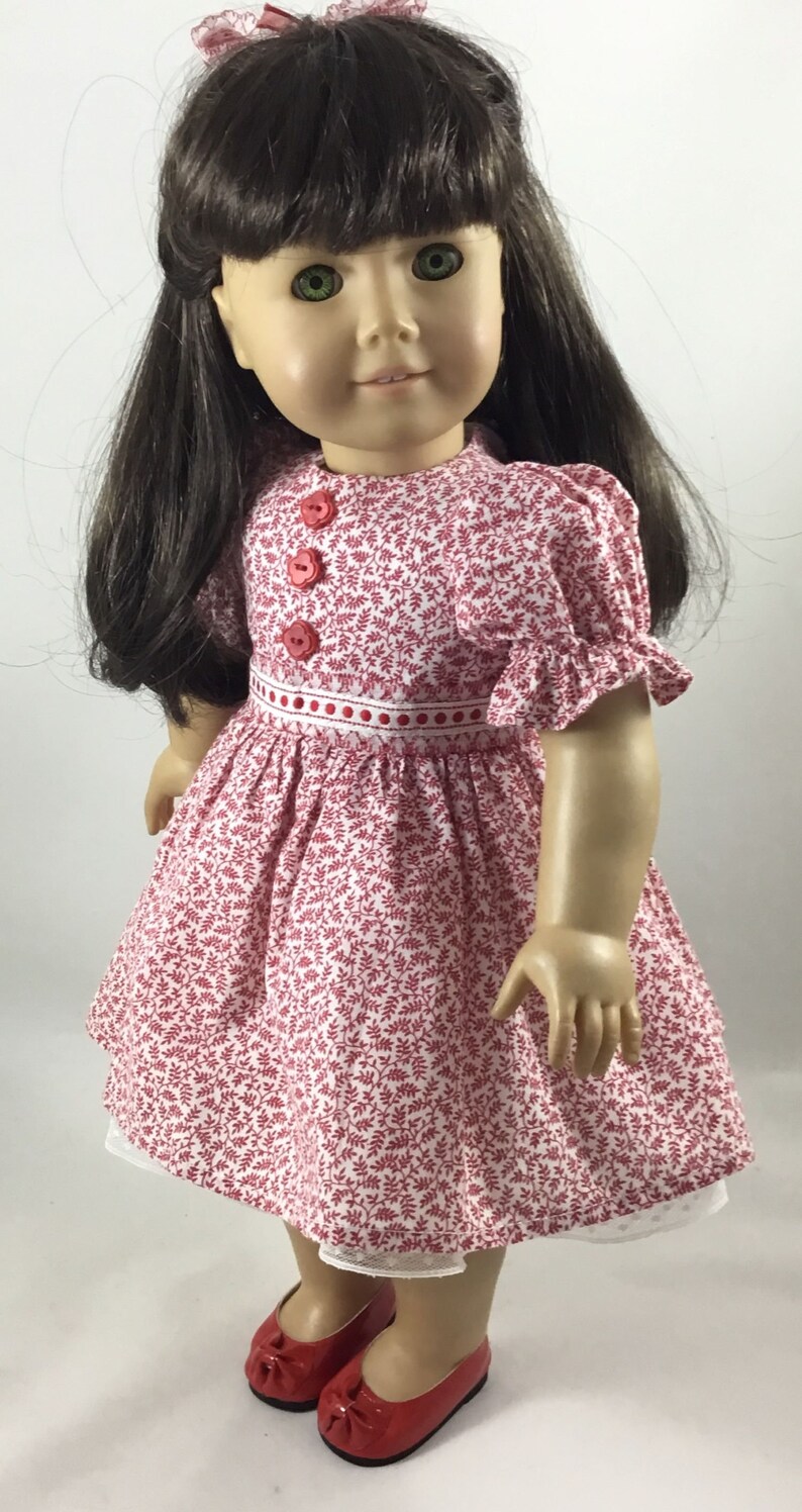 May include: A doll wearing a red and white floral dress with red buttons and a white lace trim. The doll has long brown hair and green eyes. She is wearing red shoes.