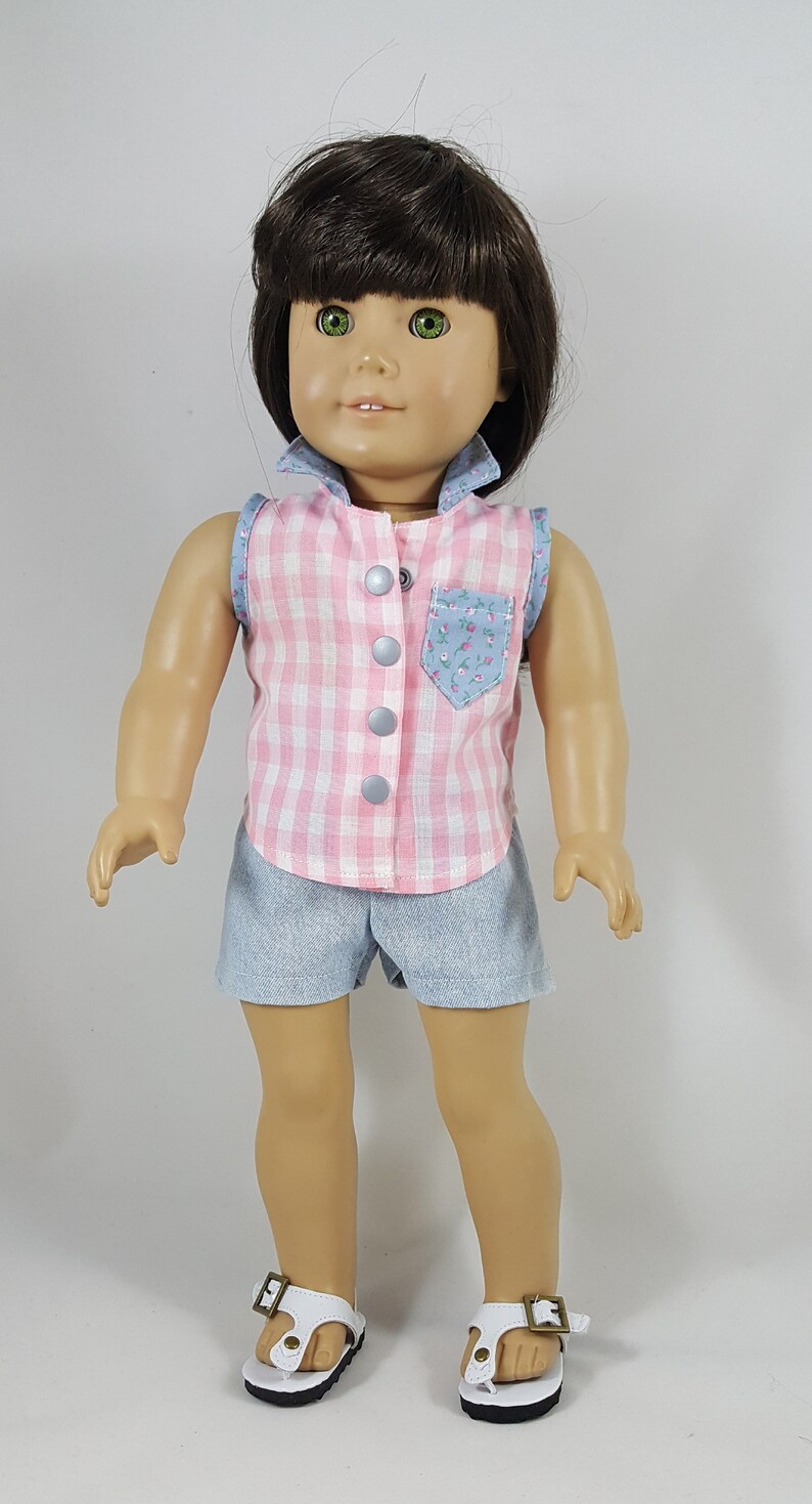 Peut inclure: Une poup&eacute;e portant une chemise sans manches en vichy rose et blanc avec une poche fleurie, un short en jean bleu et des sandales blanches &agrave; boucles.
