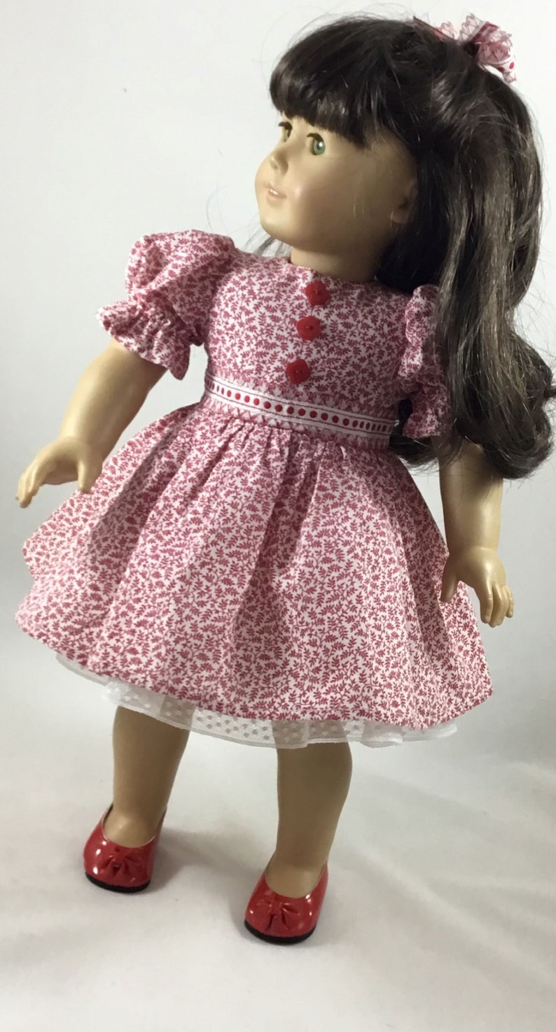 May include: A doll wearing a red and white floral dress with red buttons and a white lace petticoat. The doll has brown hair and green eyes and is wearing red shoes.